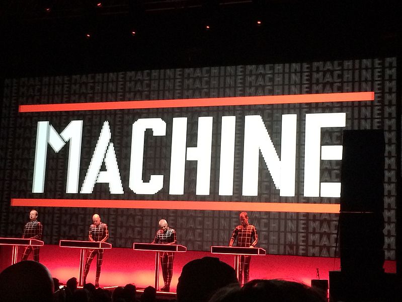 German electronic music pioneers Kraftwerk perform their signature 3D show at the Riviera Theatre in Chicago on March 27, 2014. The band members are silhouetted against a black backdrop with the word "MACHINE" repeated in multiple lines of different sizes and colors.