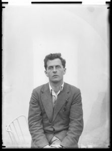A black and white portrait of Ludwig Wittgenstein (1889-1951), Austrian philosopher, logician, and mathematician. He appears pensive, gazing off to his right with intense focus. Wittgenstein has dark hair, is dressed in a buttoned-up shirt and a tweed jacket with a pocket square, and sits with his hands folded in his lap. The background is overexposed, giving his figure a slight halo effect. A white chair is partially visible to his left.